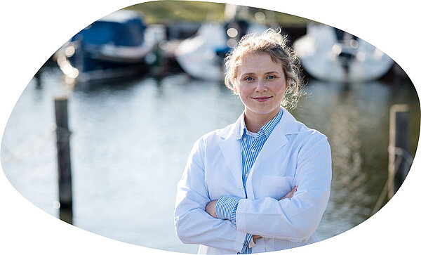 Eine lächelnde Frau in weißem Arztkittel steht mit verschränkten Armen vor einem Wasserbecken mit mehreren angelegten Booten im Hintergrund.