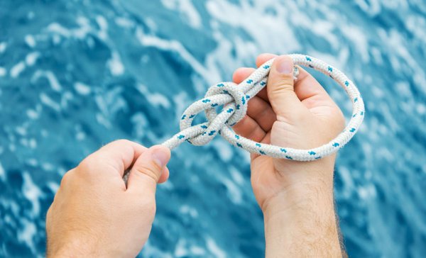 Zwei Hände knüpfen einen Seemannsknoten vor blauem Wasser