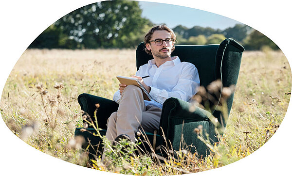 Ein Mann mit Brille sitzt in einem dunkelgrünen Sessel auf einer Wiese und schreibt in einen Notizblock. Im Hintergrund sind hohe Gräser und Bäume unter einem hellen Himmel zu sehen.