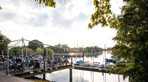 Menschen sitzen auf einer Außenterrasse an einem Fluss mit Segelbooten im Hintergrund